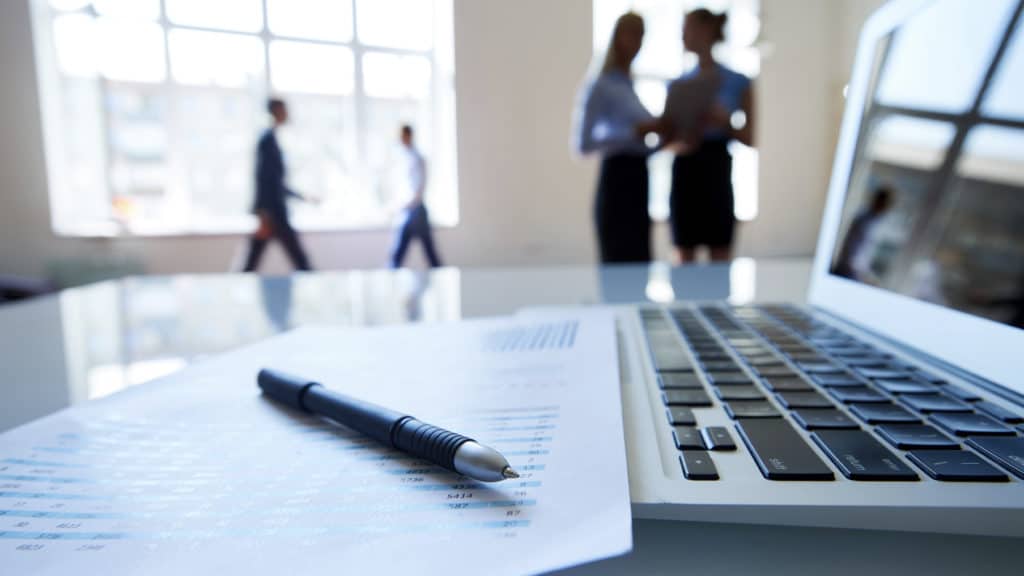 Document with financial analysis, ballpoint pen and laptop on foreground, business people in the background