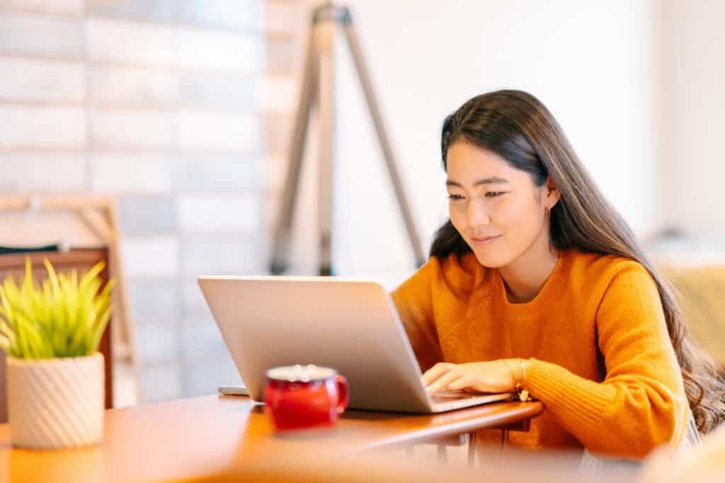 A young Asian woman wearing a bright orange shirt is using a laptop and smiling.