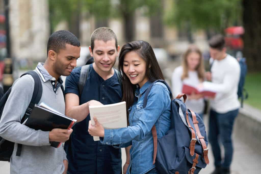 A group of students are standing outside. They are looking at a book together and appear happy.