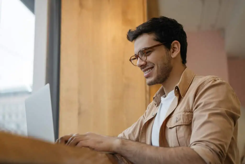 A man wearing glasses and a yellow short sleeved shirt is typing on a laptop while smiling. There is a wood panel door in the background.