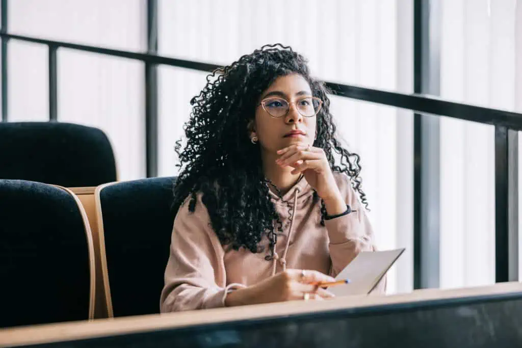 A woman with curly hair and glasses sits attentively in a lecture hall. She is holding notes.