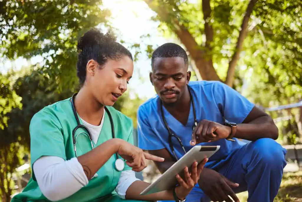 Two nurses look at a chart together with green trees in the background.