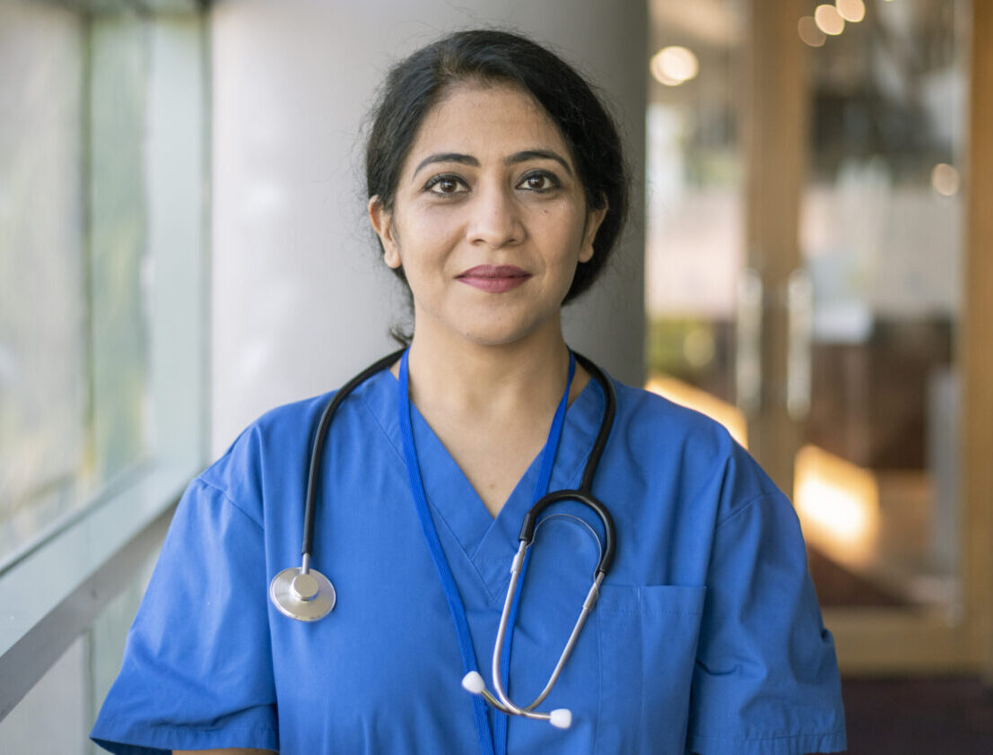 A female doctor or nurse of Indian descent wearing scrubs and a stethoscope poses next to a window in a hospital corridor and smiles confidently directly at the camera.